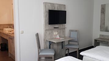 Una habitación de hotel compacta con una cama, un escritorio con silla, un televisor montado en la pared, una mesita y un espejo.