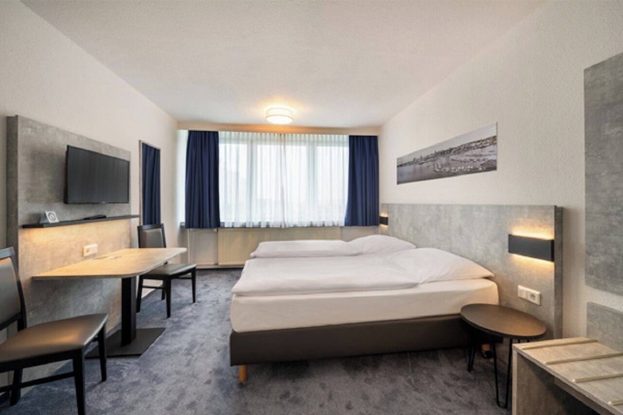 Habitación de hotel con cama, escritorio, silla, mesita, televisor montado en la pared y ventana con cortinas.