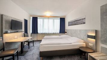 Una camera d'albergo con un letto, una scrivania, una sedia, un tavolino, una TV montata a parete e una finestra con tende.