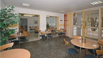 A room with multiple tables and chairs, a large plant, and a view through a window with yellow vertical blinds.
