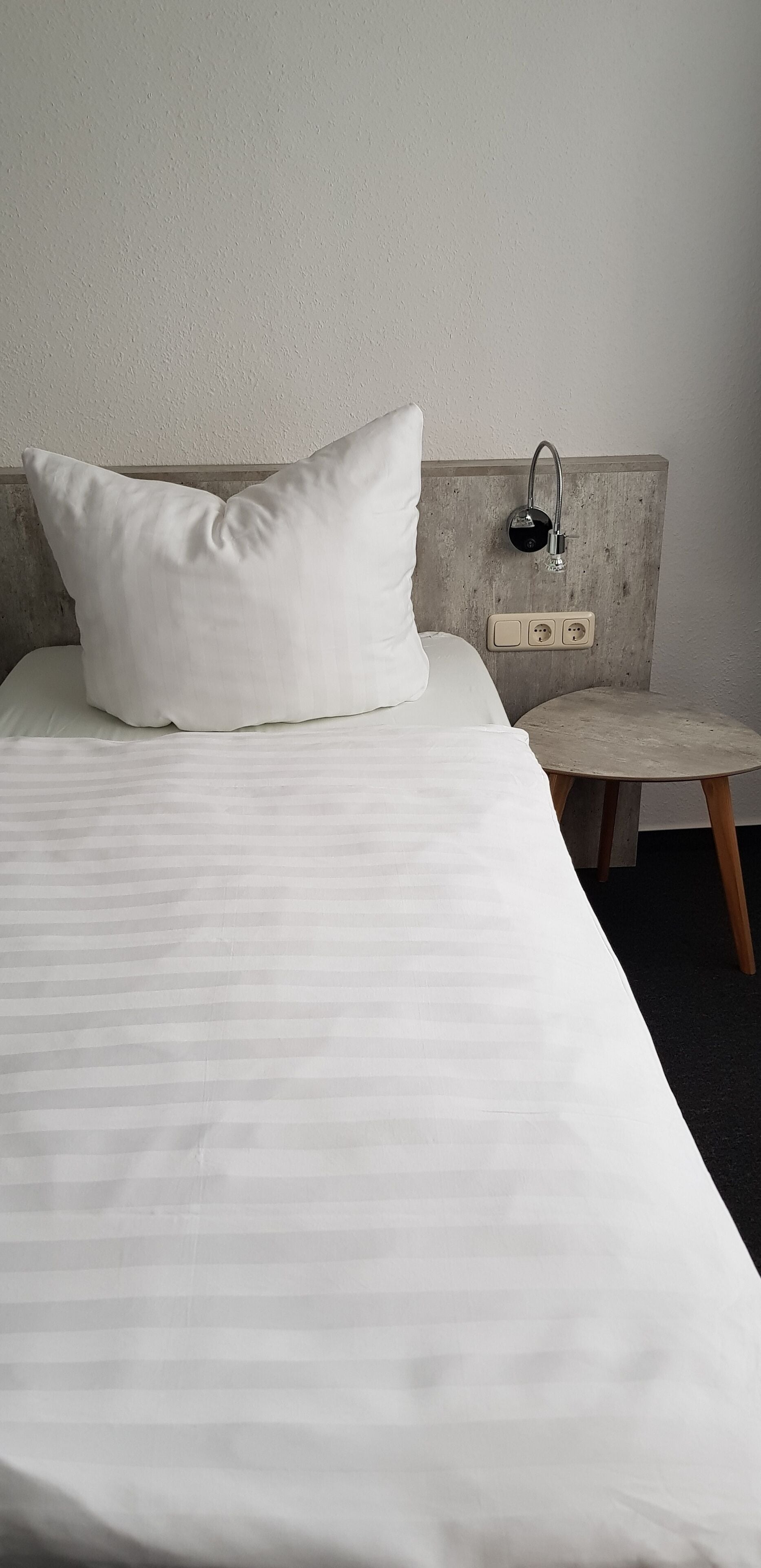 A neatly made bed with a pillow, a small round table, and a wall-mounted light fixture.