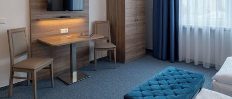 Een hotelkamer met een houten bureau, twee stoelen, een televisie, een kledingkast, een blauwe poef en een bed.