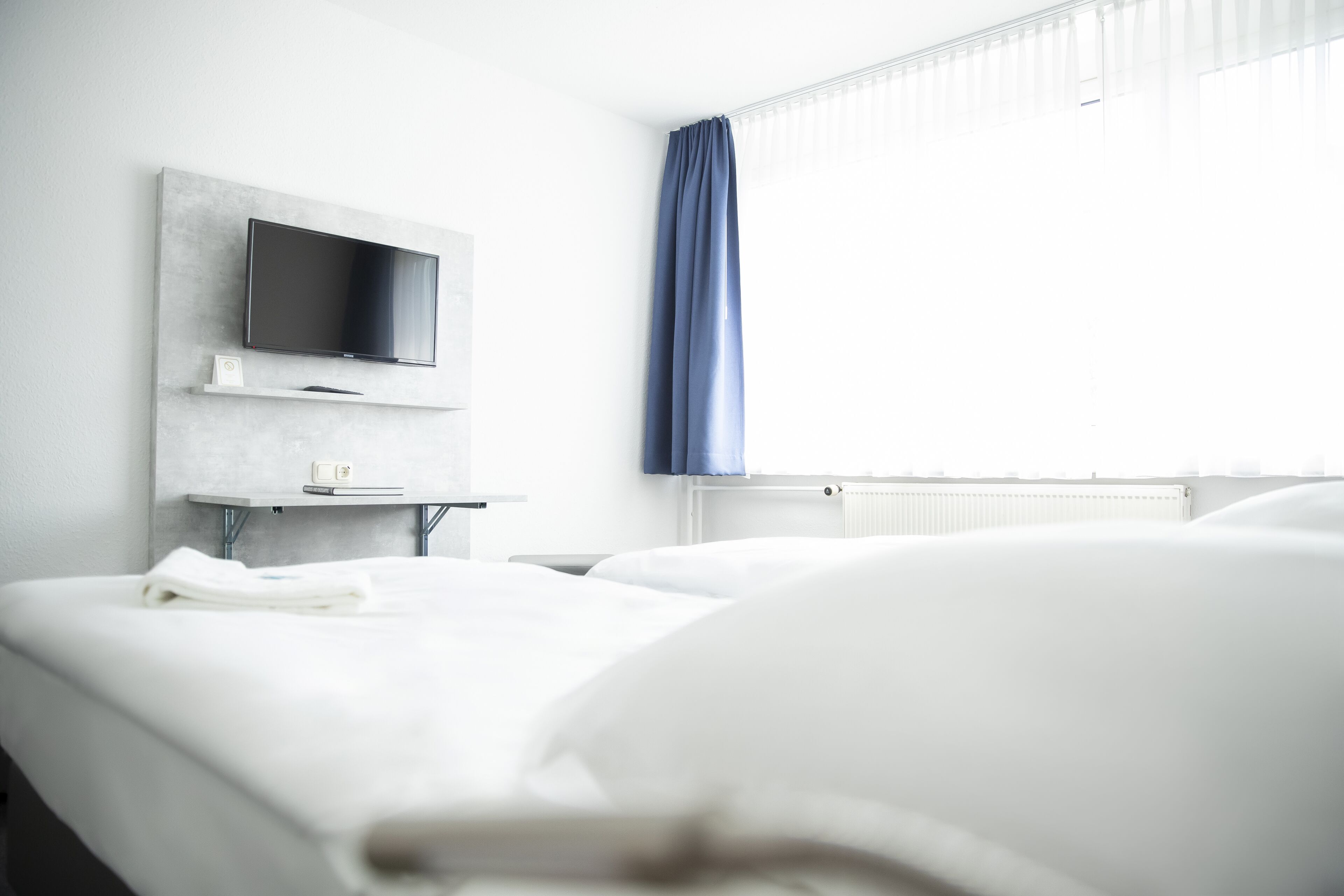 Habitació d'hotel amb un llit, un televisor de pantalla plana muntat a una paret gris, una taula petita amb una làmpada i una finestra amb cortines blaves.