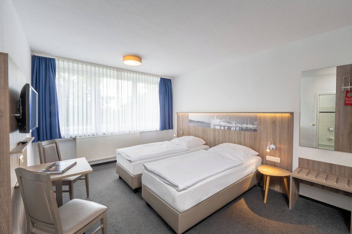 Habitación de hotel con dos camas, un escritorio, una silla, un televisor y un ventanal con cortinas.