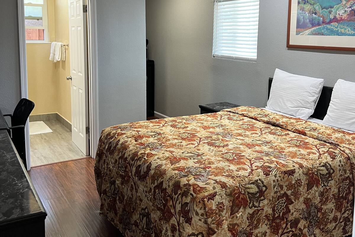 A bedroom with a floral bedspread, a desk, a chair, a window with blinds, and a framed landscape painting on the wall.