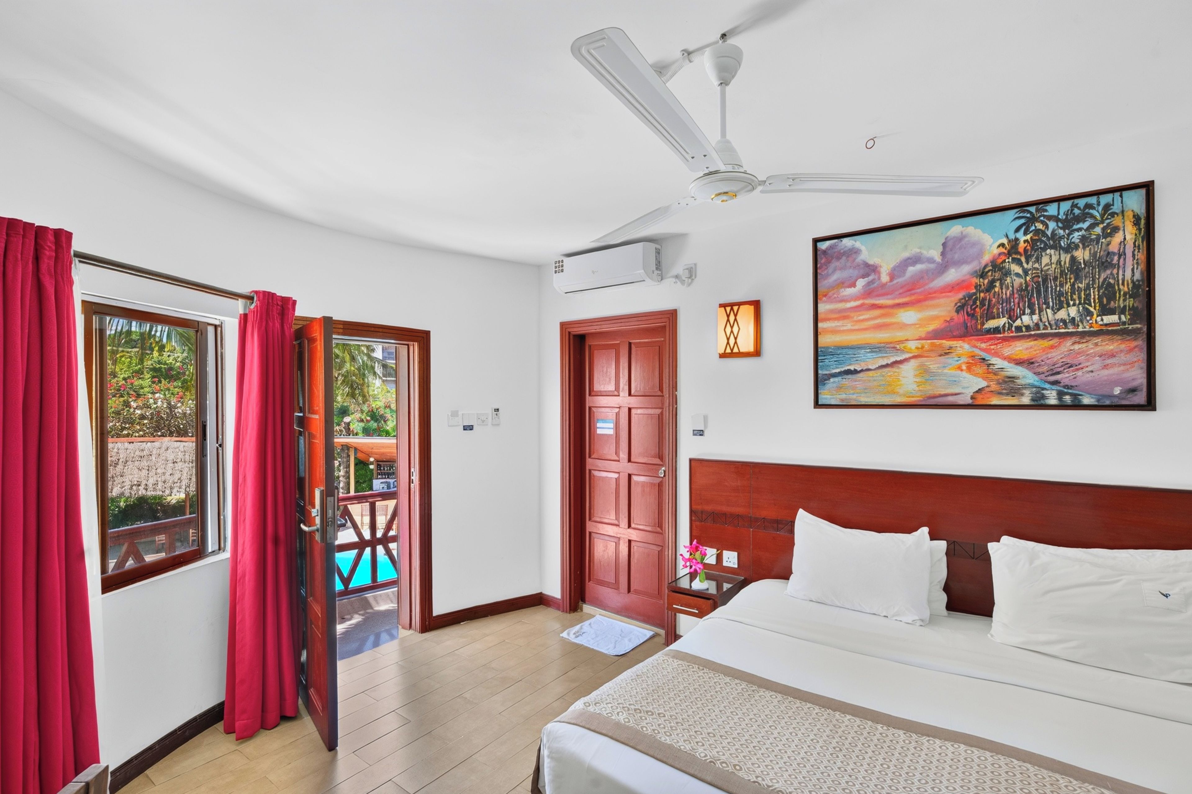 Ein Hotelzimmer mit Bett, Deckenventilator, einem Gemälde und Blick auf Pool und Garten.