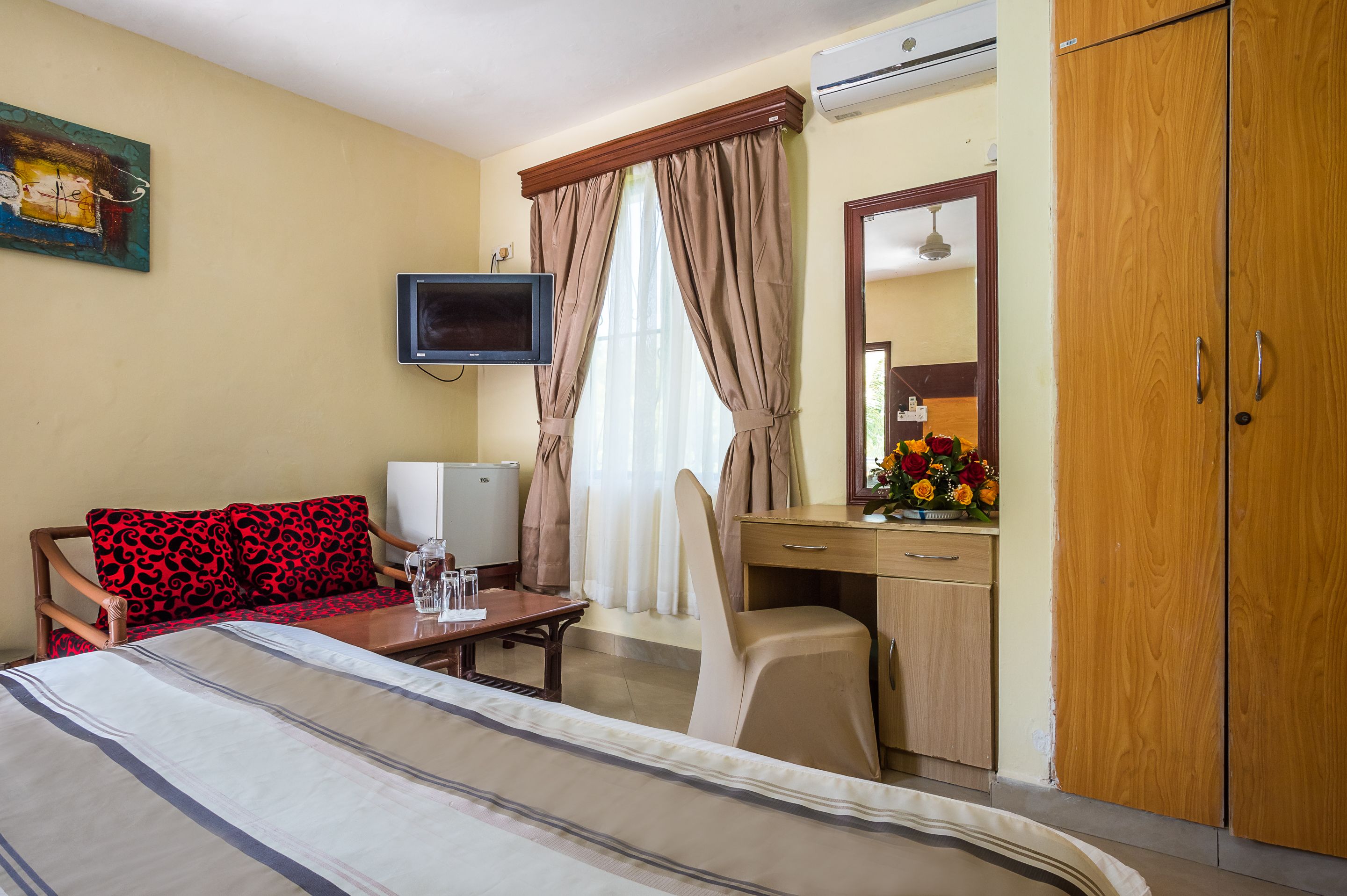 Quarto de hotel com cama, escrivaninha com cadeira, televisão, guarda-roupa e janela com cortinas.