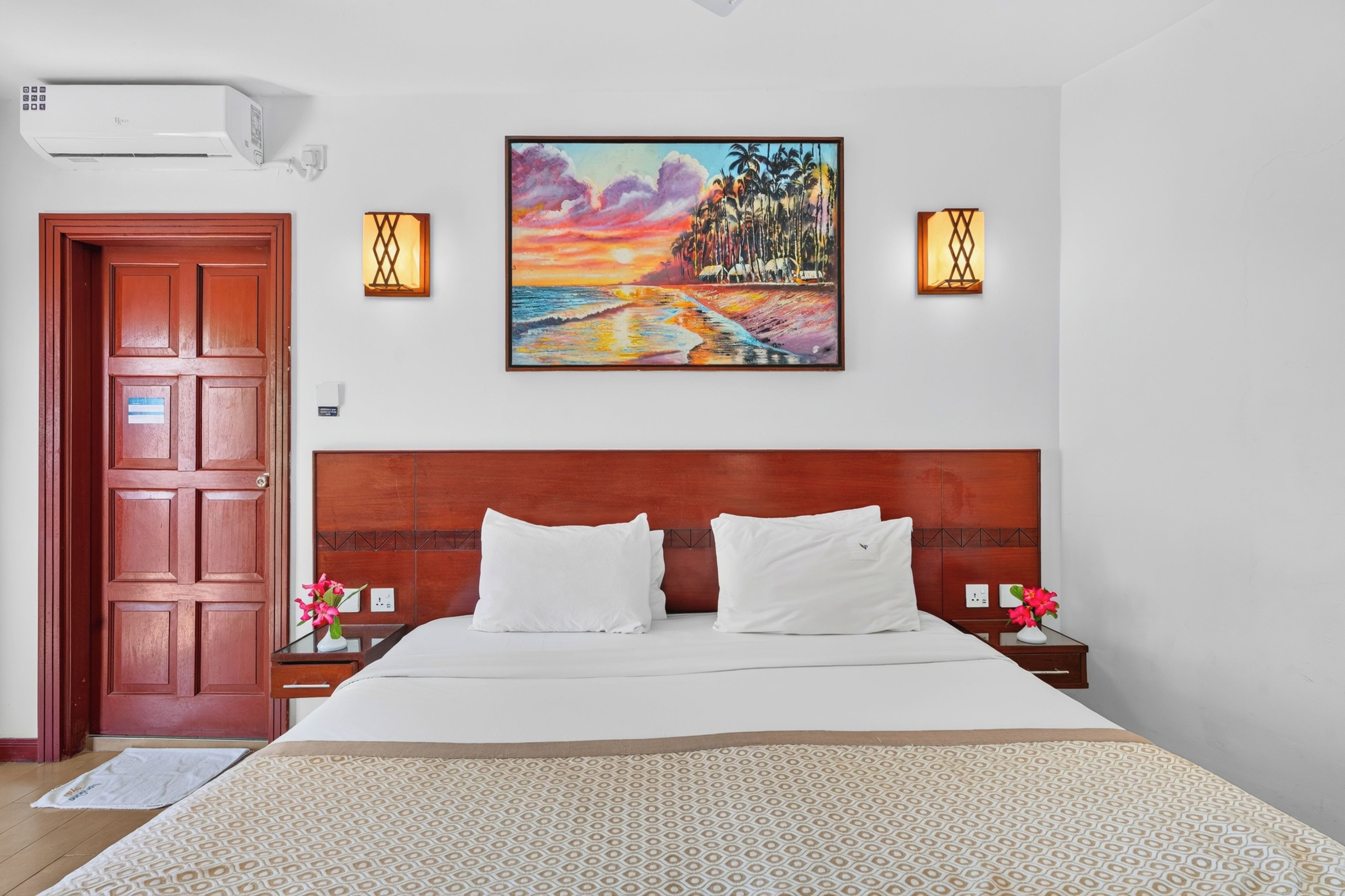 A hotel room with a wooden headboard, a large bed with white bedding, two bedside tables with lamps, and a painting of a sunset over a beach.