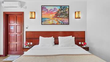 A hotel room with a wooden headboard, a large bed with white bedding, two bedside tables with lamps, and a painting of a sunset over a beach.