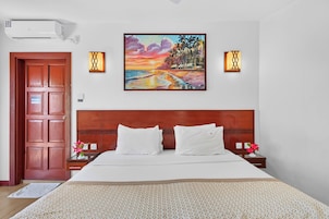 Habitación de hotel con cabecera de madera, una cama grande con ropa blanca, dos mesitas de noche con lámparas y un cuadro de un atardecer sobre la playa.