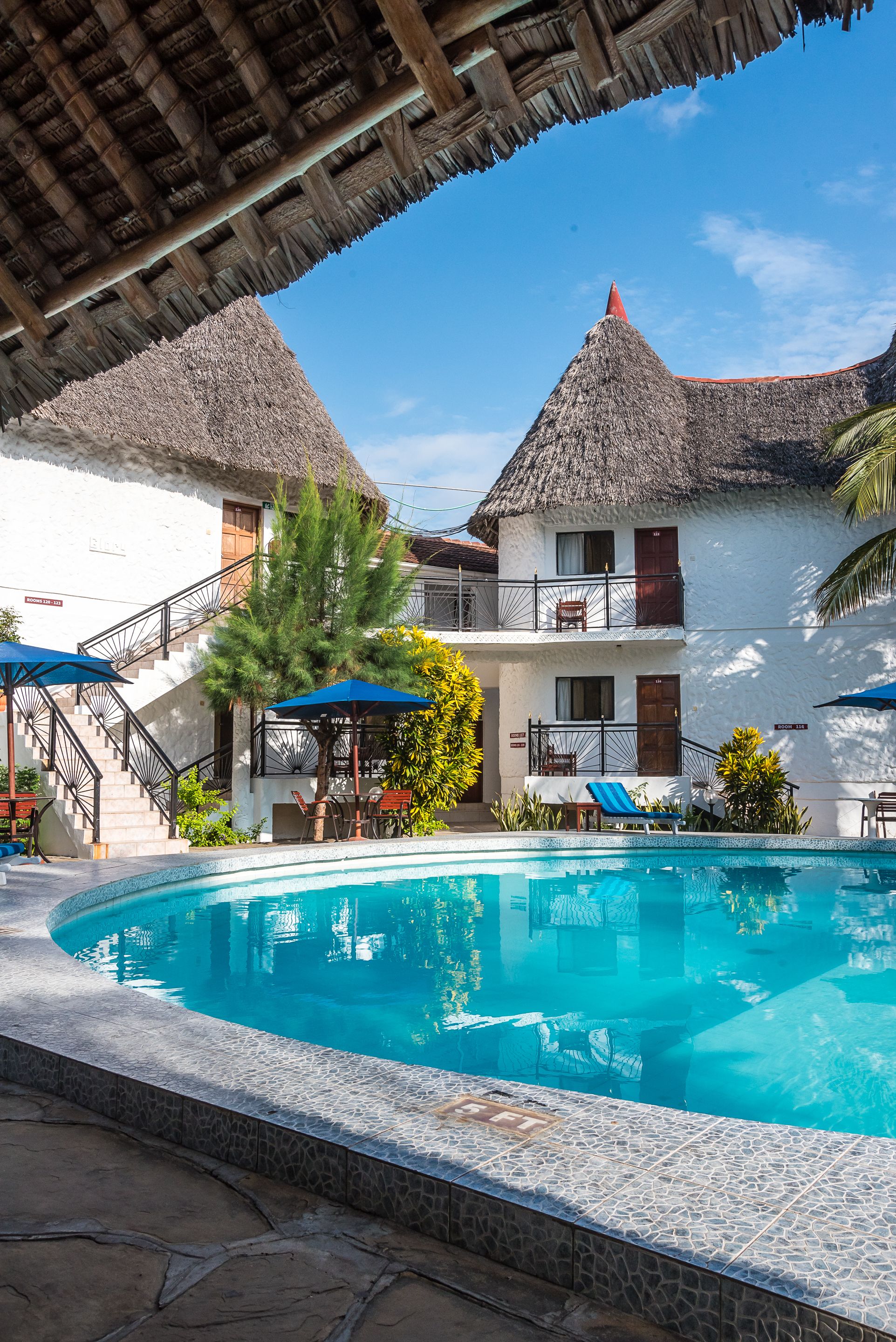 A pool area with a clear blue pool, a staircase leading to a balcony, and a thatched roof structure.