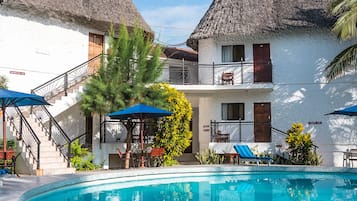 Un área de piscina con una piscina azul cristalina, una escalera que lleva a un balcón y una estructura con techo de paja.
