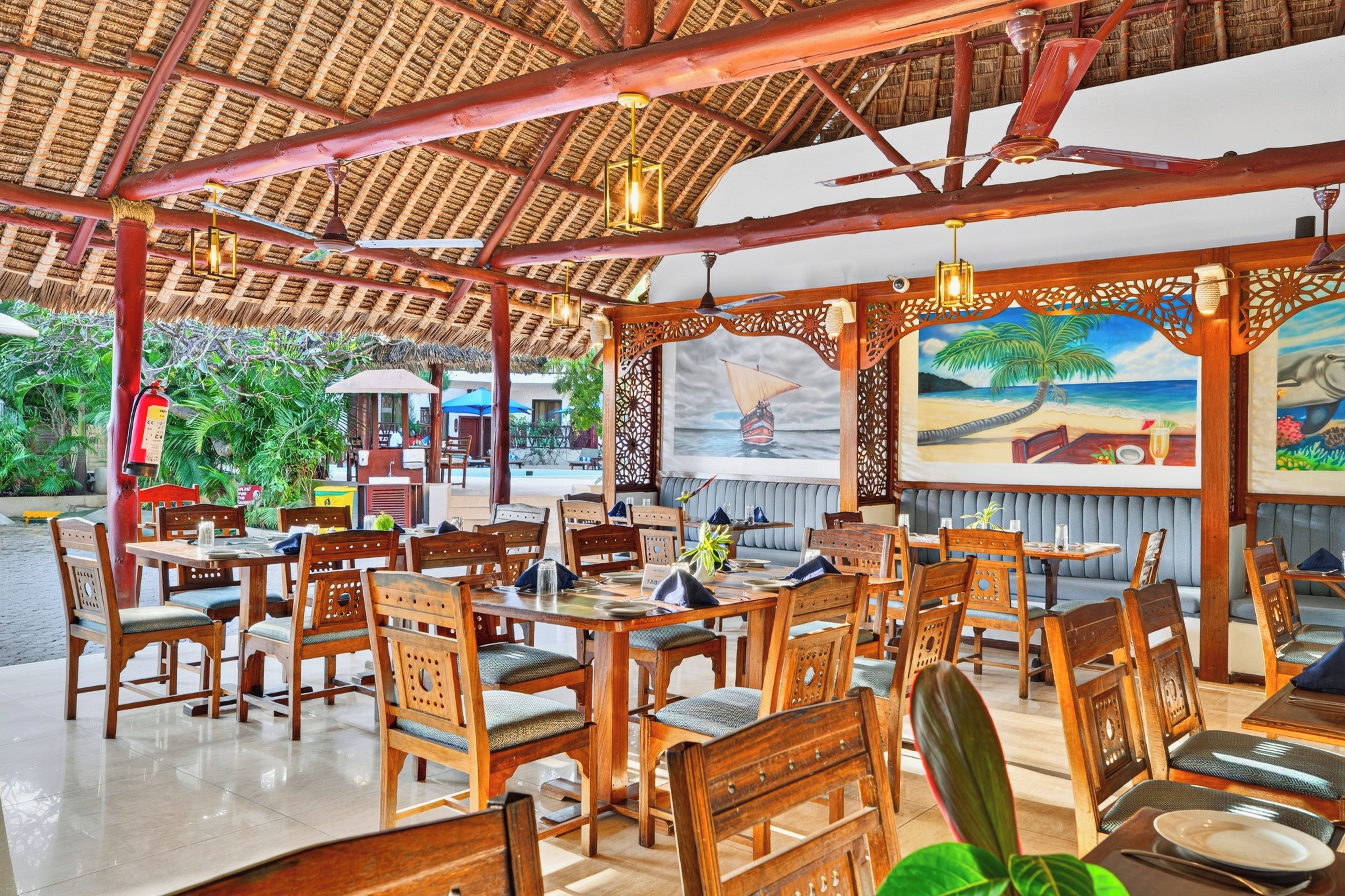 Ein Restaurant im Freien mit Holztischen und -stühlen, einem Reetdach und einer Wandmalerei mit Strandmotiv.