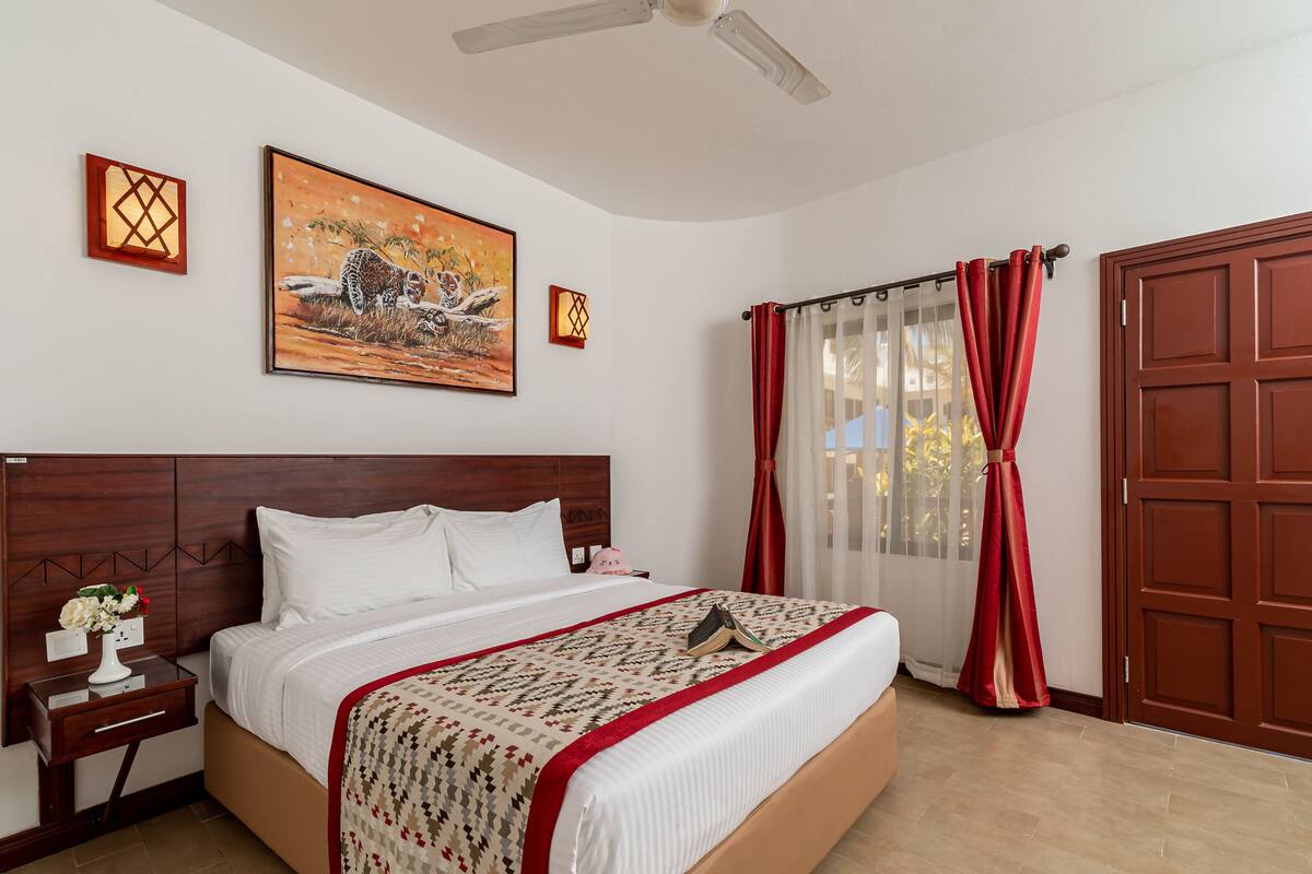 A hotel room with a large bed, wooden headboard, a nightstand with a vase of flowers, a painting on the wall, and a window with red curtains.