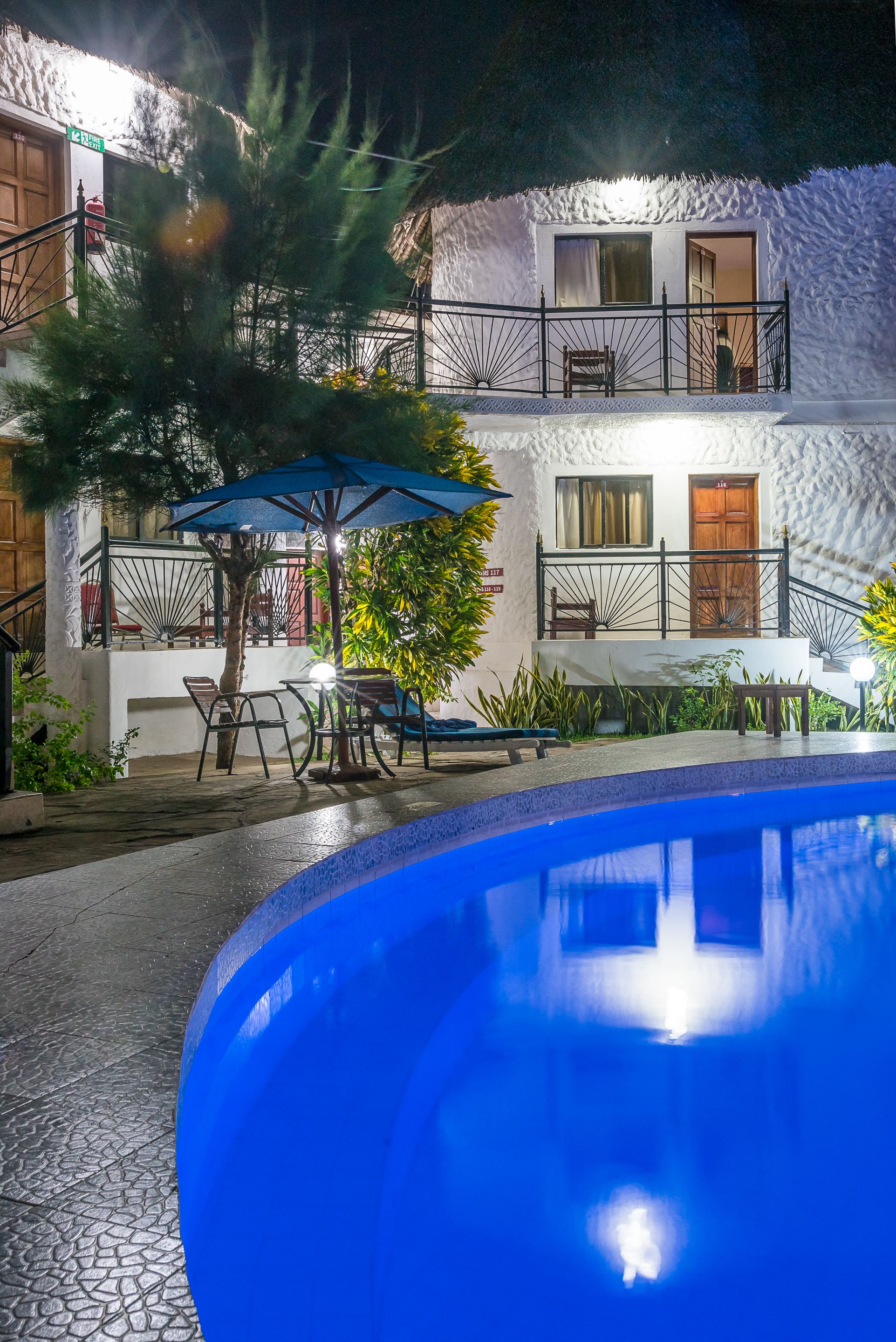 A poolside area with a white building, outdoor seating, and a staircase leading to a balcony.