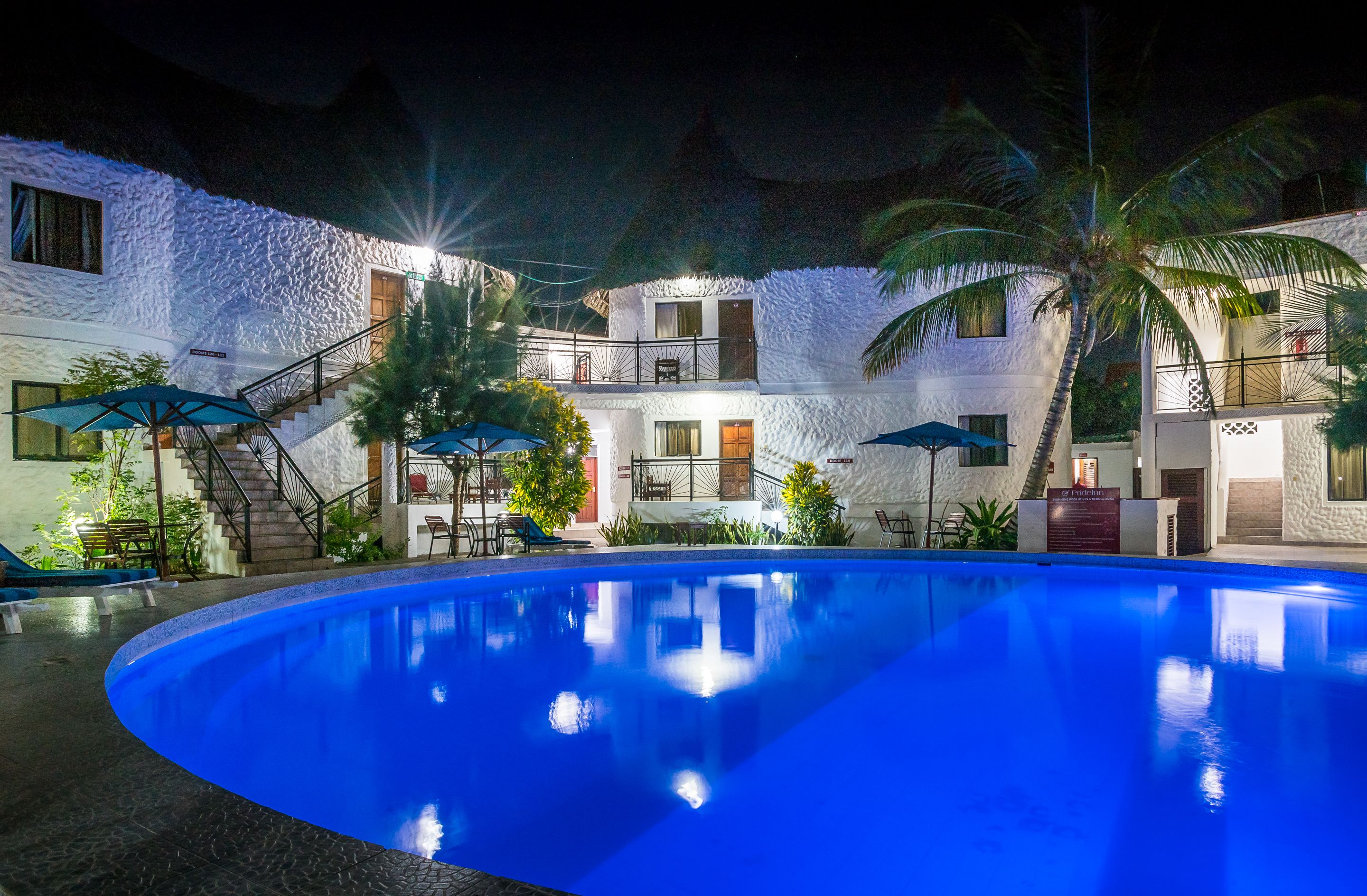 Una piscina con pared de piedra y palmeras, rodeada por un edificio con balcones y zona de estar al aire libre.