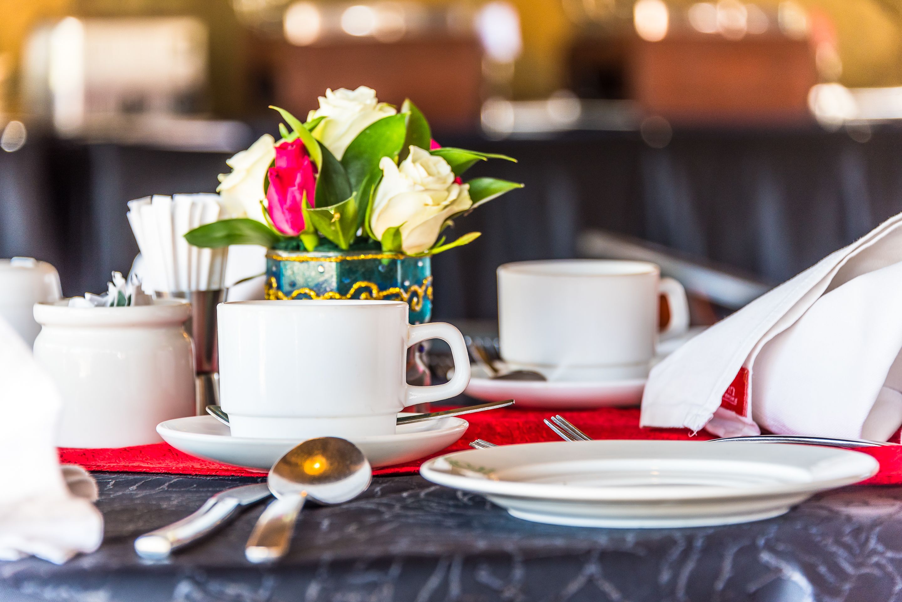 A dining table set with white cups, saucers, and a spoon, with a vase of flowers in the center.
