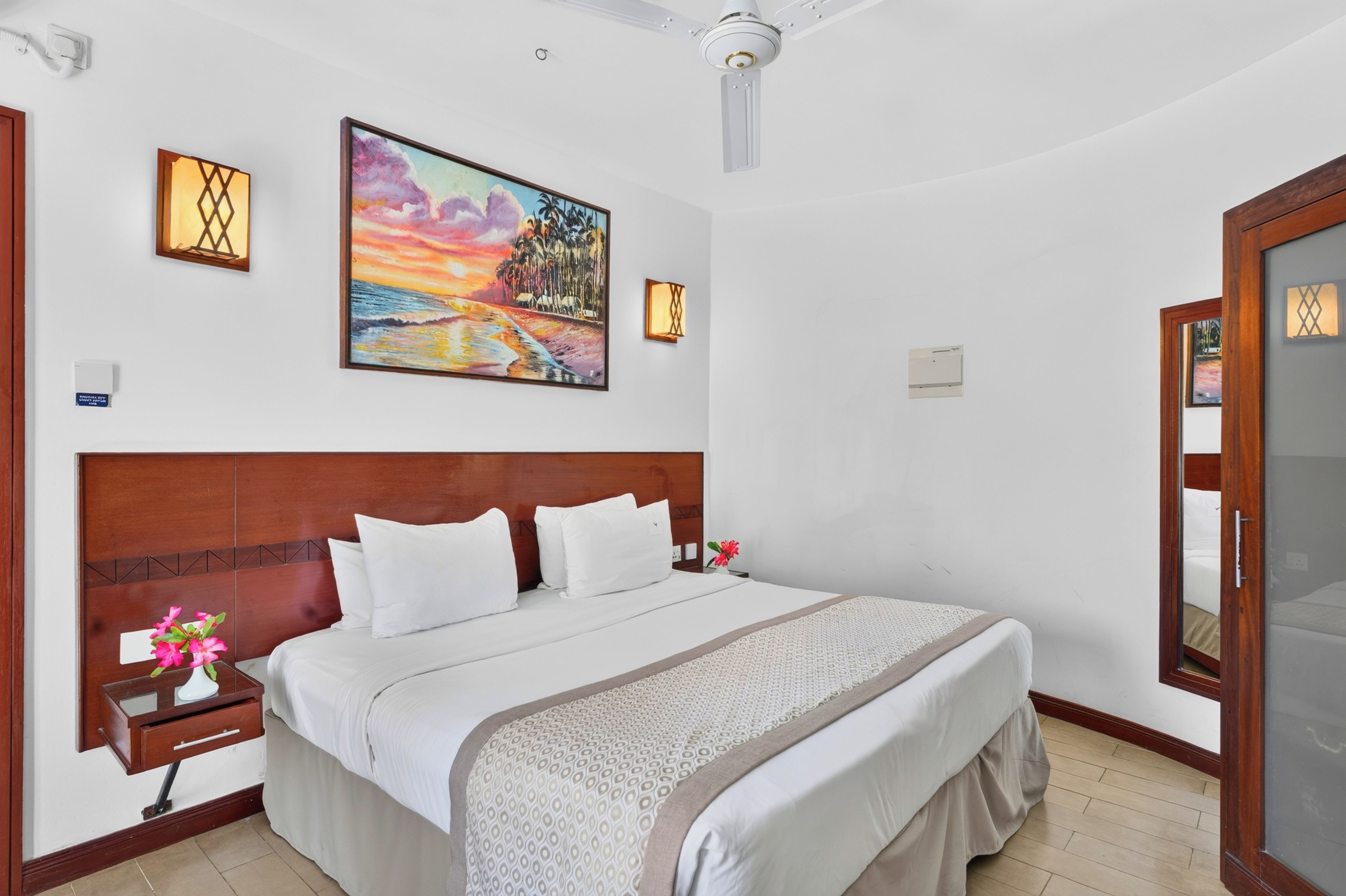 A hotel room with a large bed, a wooden headboard, a painting on the wall, and a ceiling fan.