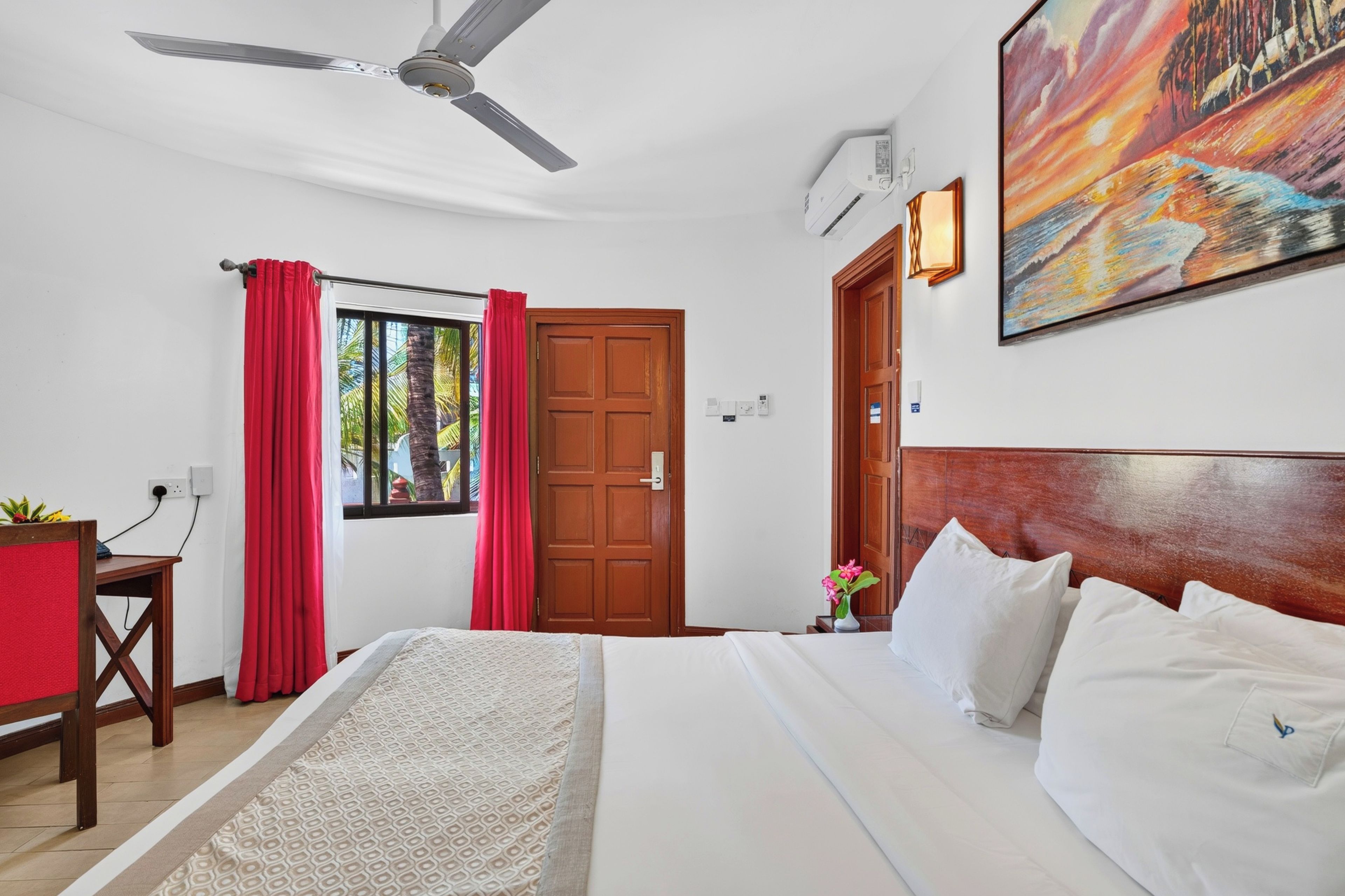 A hotel room with a bed, a desk, a ceiling fan, a door, and a painting on the wall.