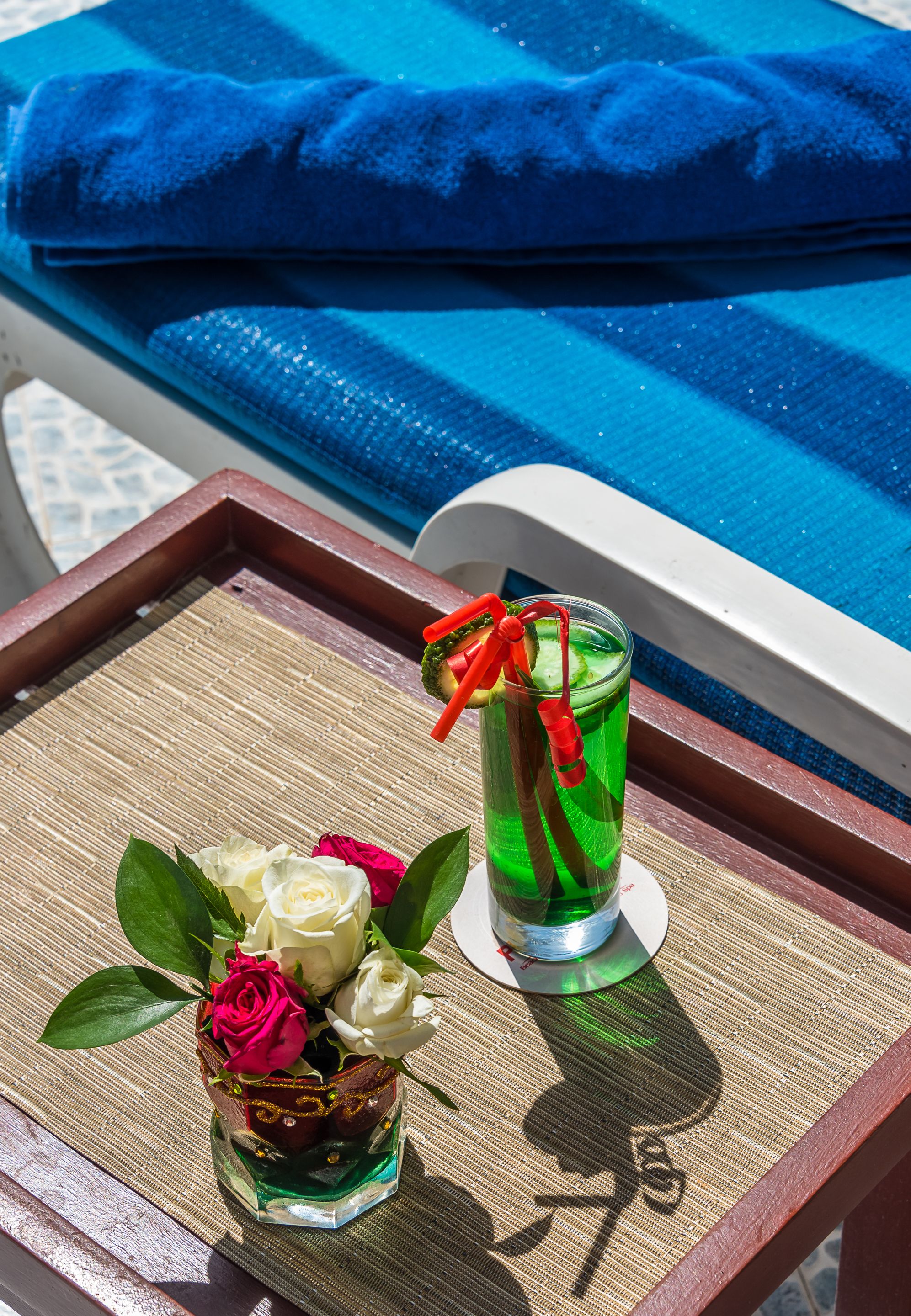 A tray with a glass of green drink and a vase of white and pink roses on a poolside lounge chair.