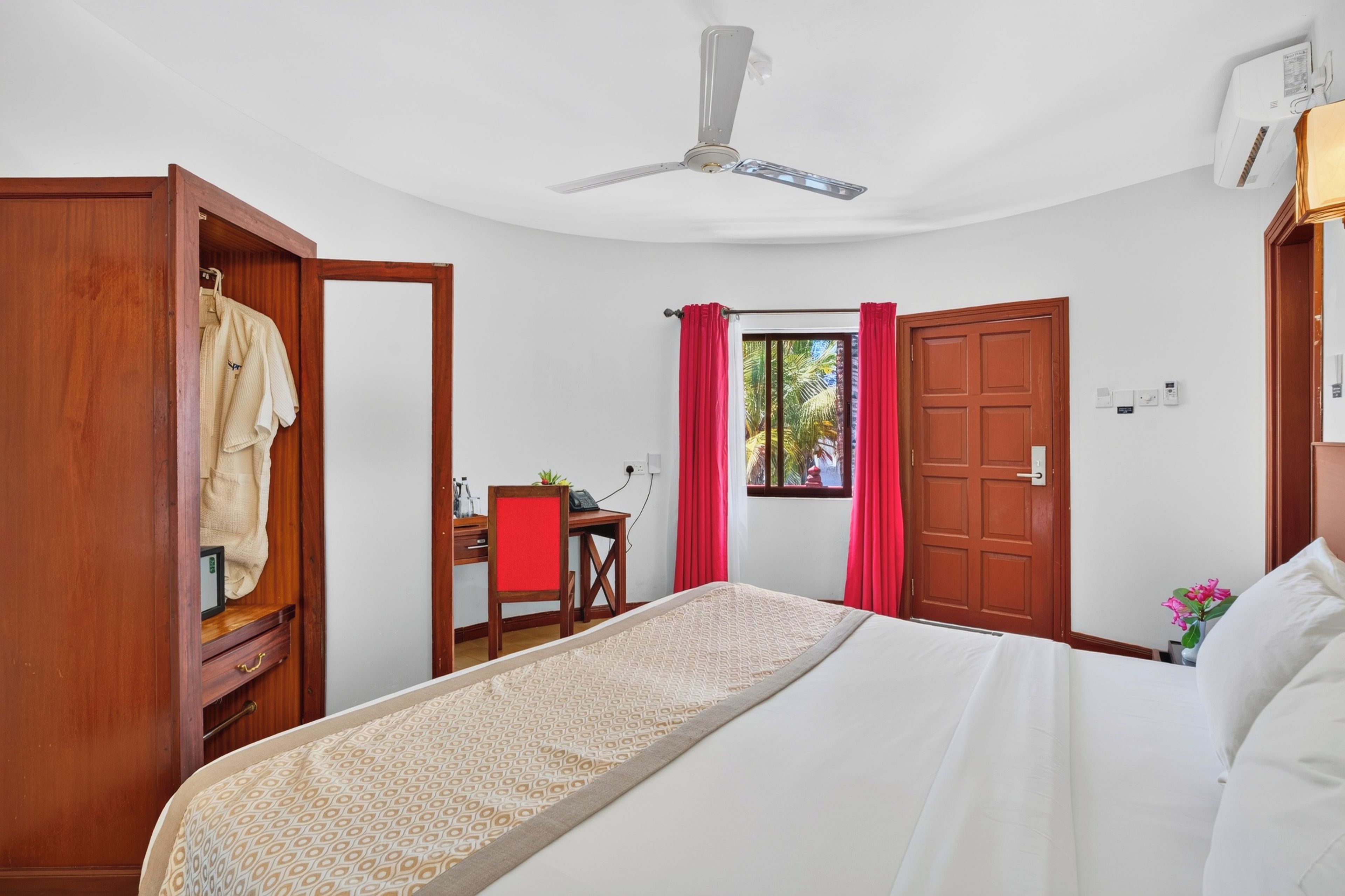 Une chambre d’hôtel équipée d’un lit, d’un bureau, d’une chaise, d’une armoire, d’un ventilateur de plafond et d’une fenêtre avec des rideaux.