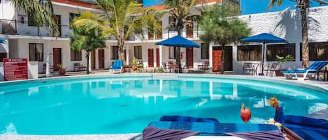 A pool area with lounge chairs and umbrellas, surrounded by palm trees and a white building with multiple windows and doors.
