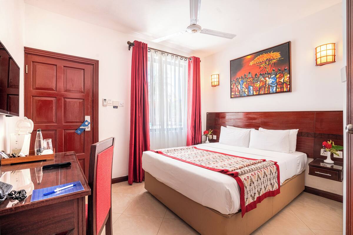 A hotel room with a large bed, a desk with a telephone and a laptop, a ceiling fan, a painting on the wall, and a window with red curtains.