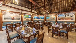 Um restaurante com móveis de madeira, mesas preparadas para refeições e murais nas paredes retratando paisagens tropicais.