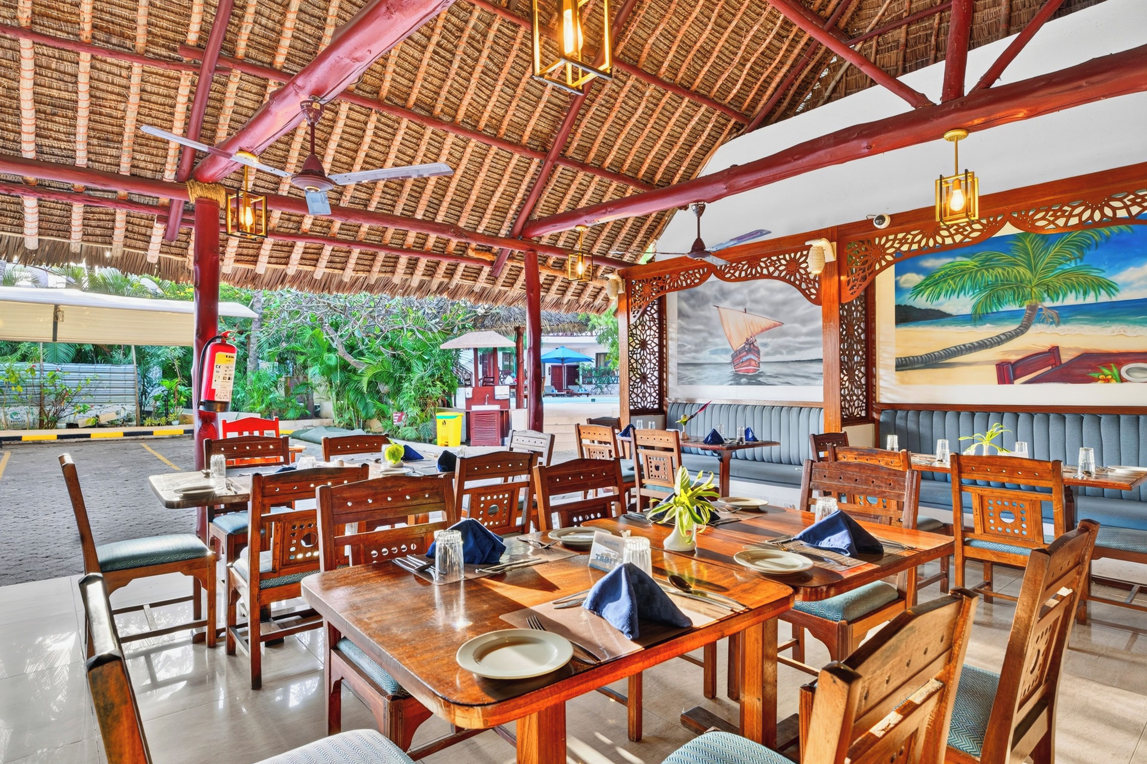 A restaurant with wooden tables and chairs, a thatched roof, and a painting of a beach scene.