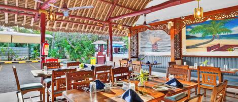 A restaurant with wooden tables and chairs, a thatched roof, and a painting of a beach scene.