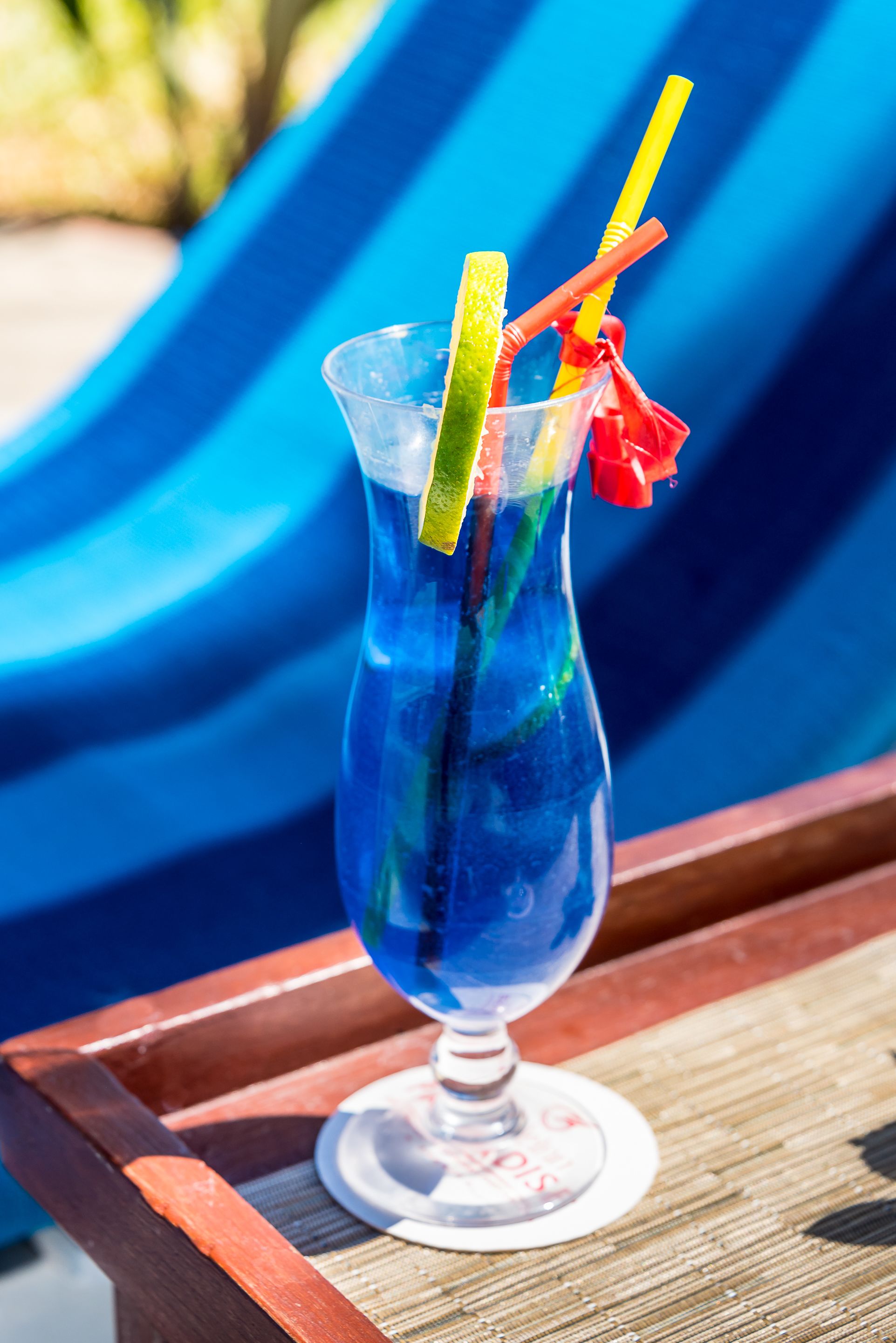 Een blauwe cocktail in een glas met een rietje en een stukje limoen op een houten dienblad.