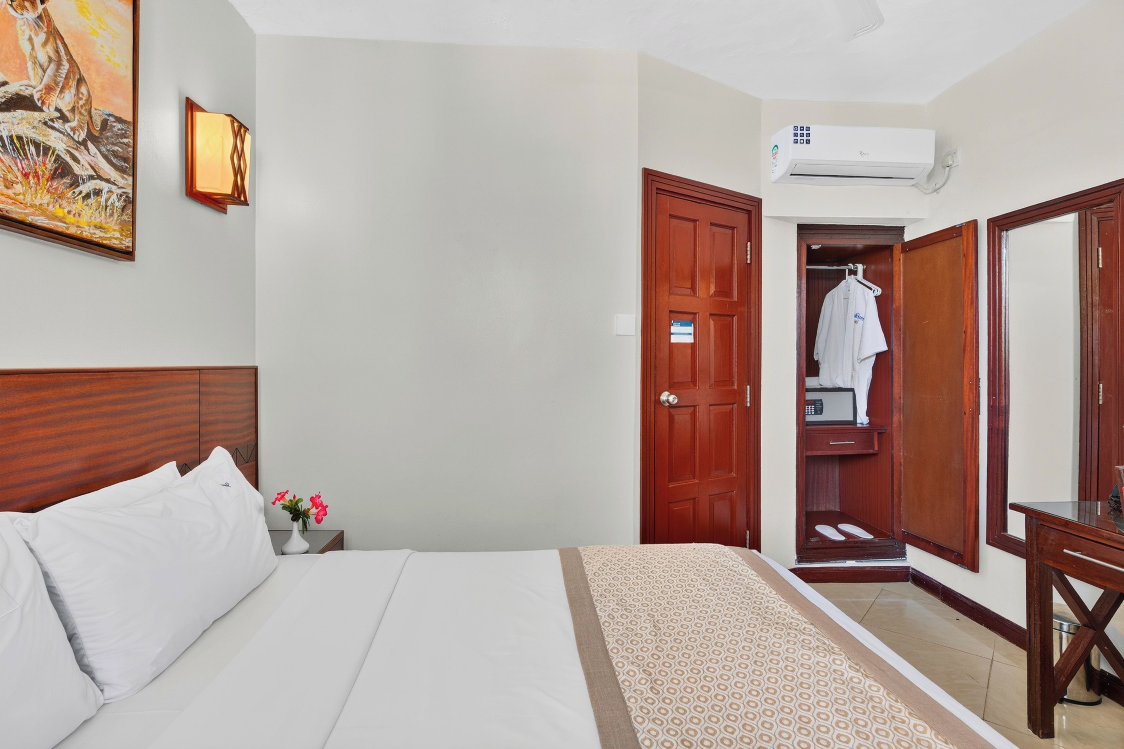 A hotel room with a bed, a nightstand, a painting, a wardrobe, and an air conditioning unit.