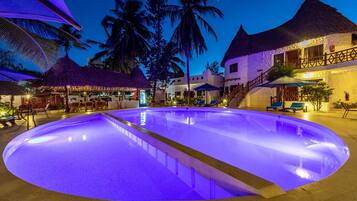 Ein Pool mit geschwungener Kante, umgeben von strohgedeckten Gebäuden und Palmen, mit einer Treppe, die zu einem Balkon führt.