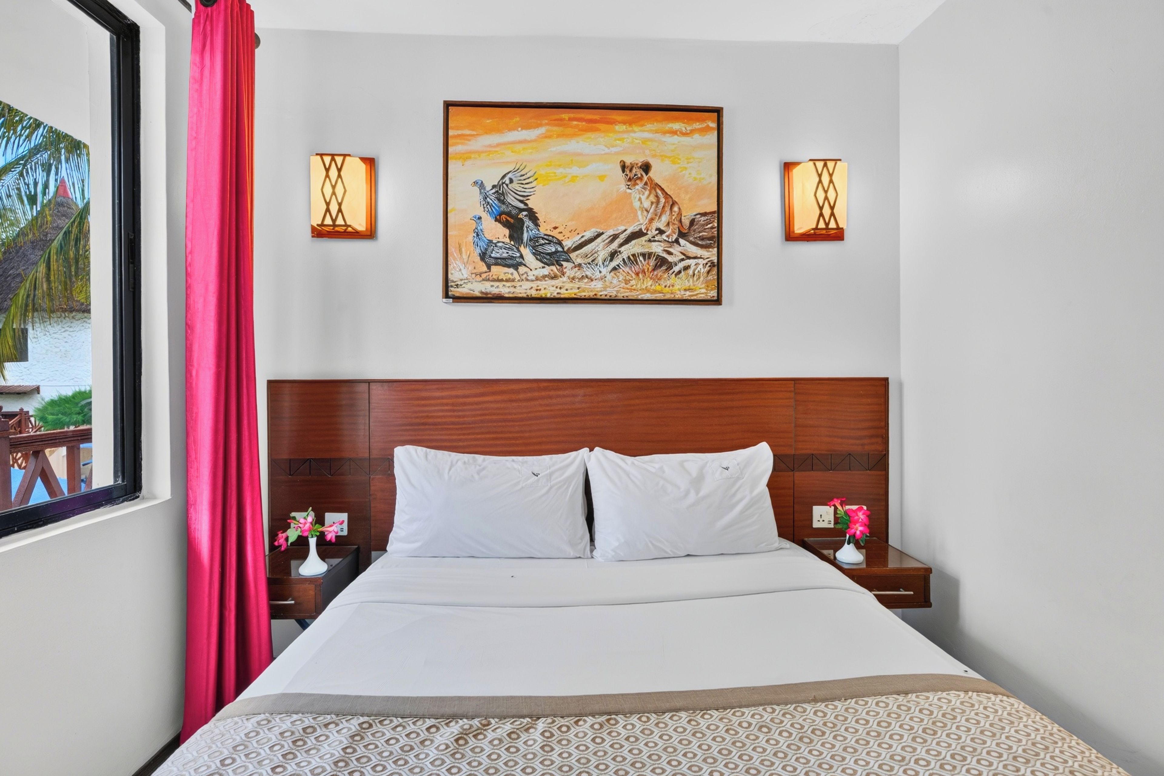 A hotel room with a bed, a painting, two wall sconces, and a window with a view of palm trees.