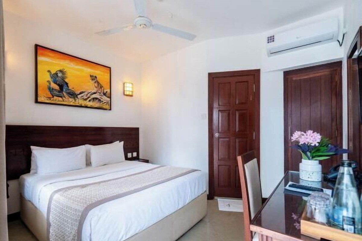A hotel room with a bed, a ceiling fan, a painting, a wooden door, and a table with a vase and bottles.