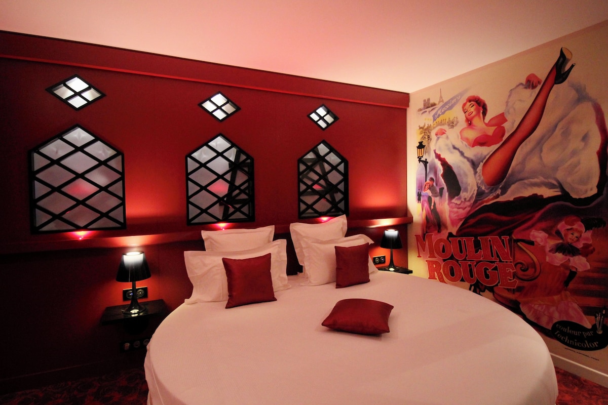 A hotel room with a large bed, red pillows, a wall with decorative windows, and a Moulin Rouge themed mural.