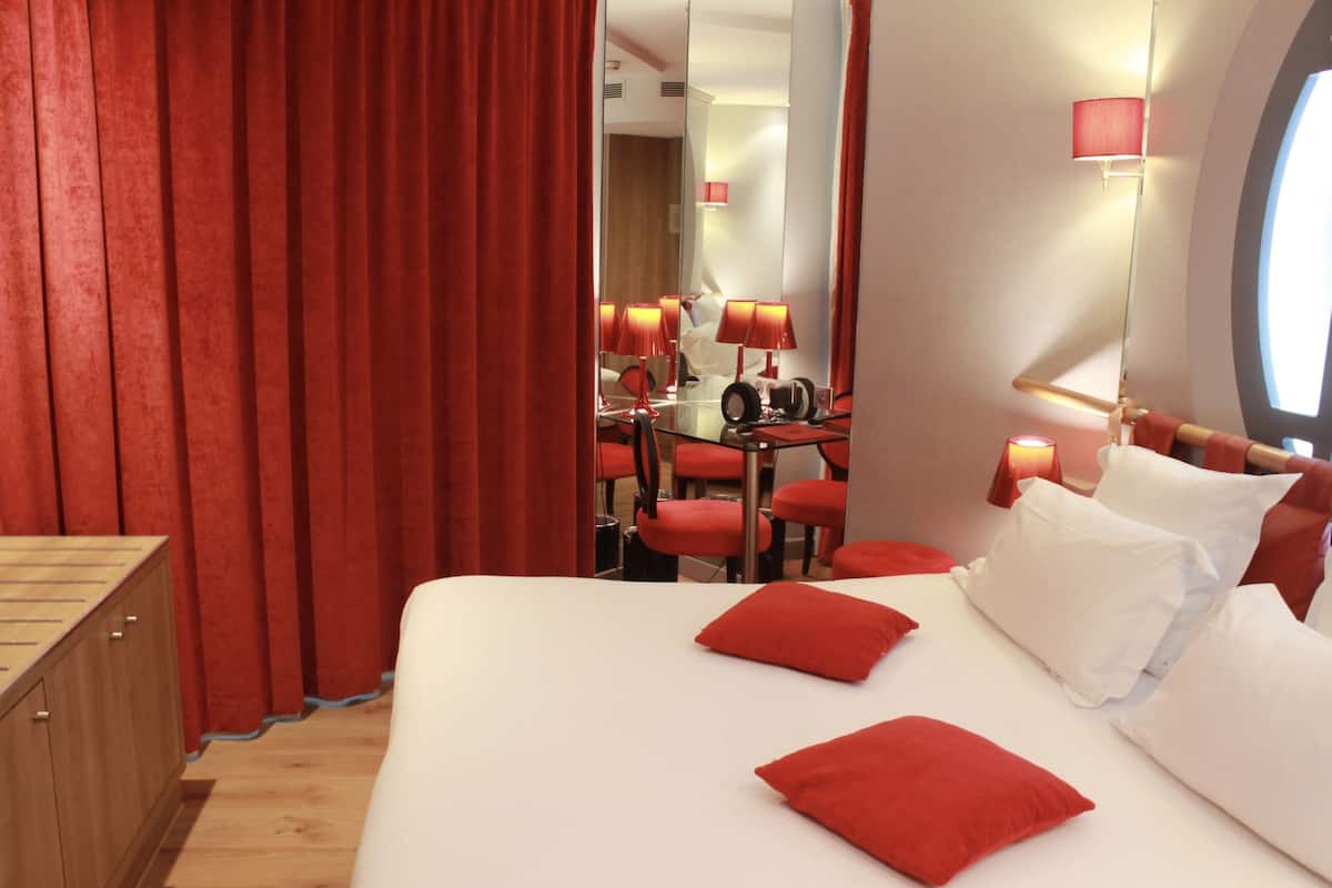 Une chambre d’hôtel avec un grand lit, des oreillers rouges, une commode en bois et un miroir qui reflète un coin repas avec des chaises et une table rouges.