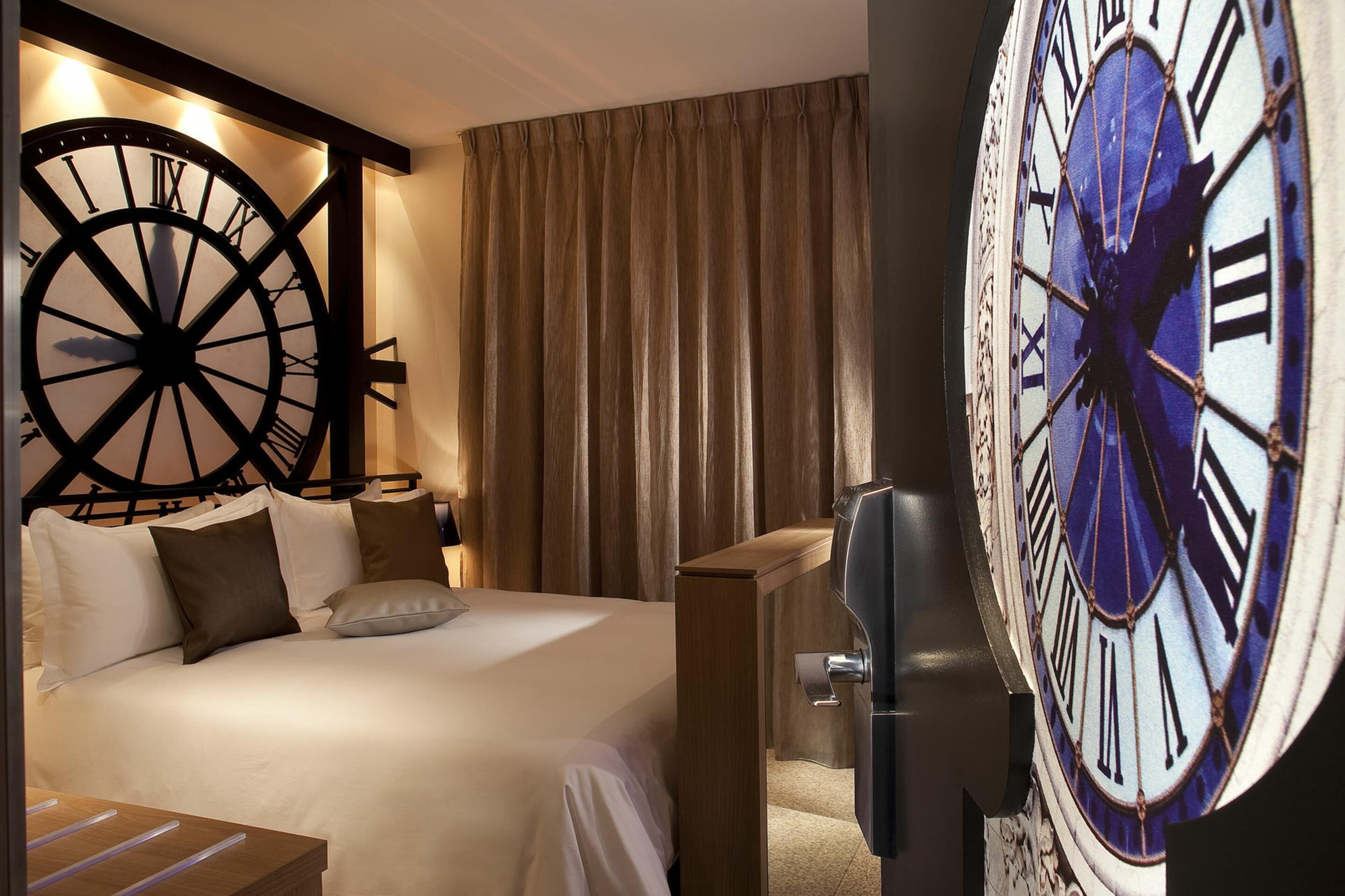 A hotel room with a large bed, a wooden nightstand, and a wall-mounted clock.