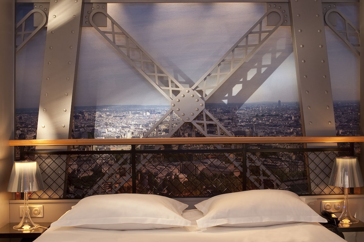Una cama con almohadas blancas y vista a un paisaje urbano a través de una ventana con estructura metálica.