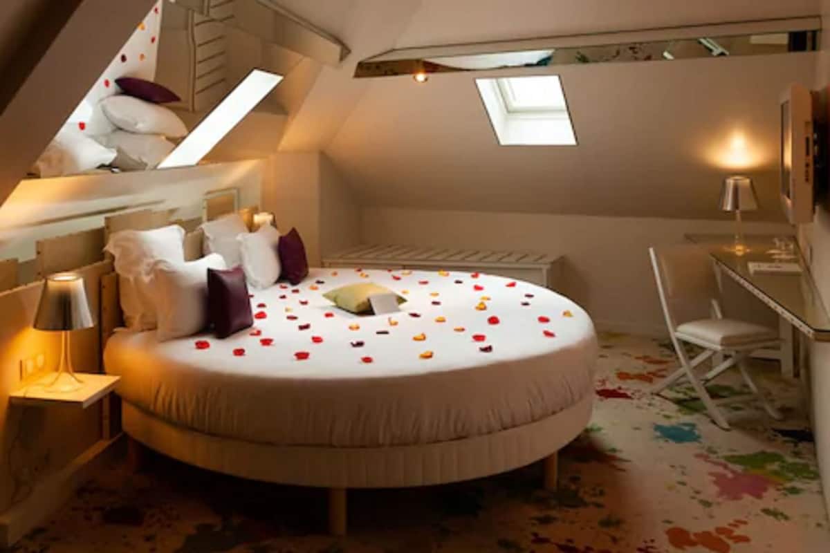 Un lit rond décoré de pétales de rose, un bureau et une chaise dans une pièce dotée d’un plafond en pente et de fenêtres de toit.