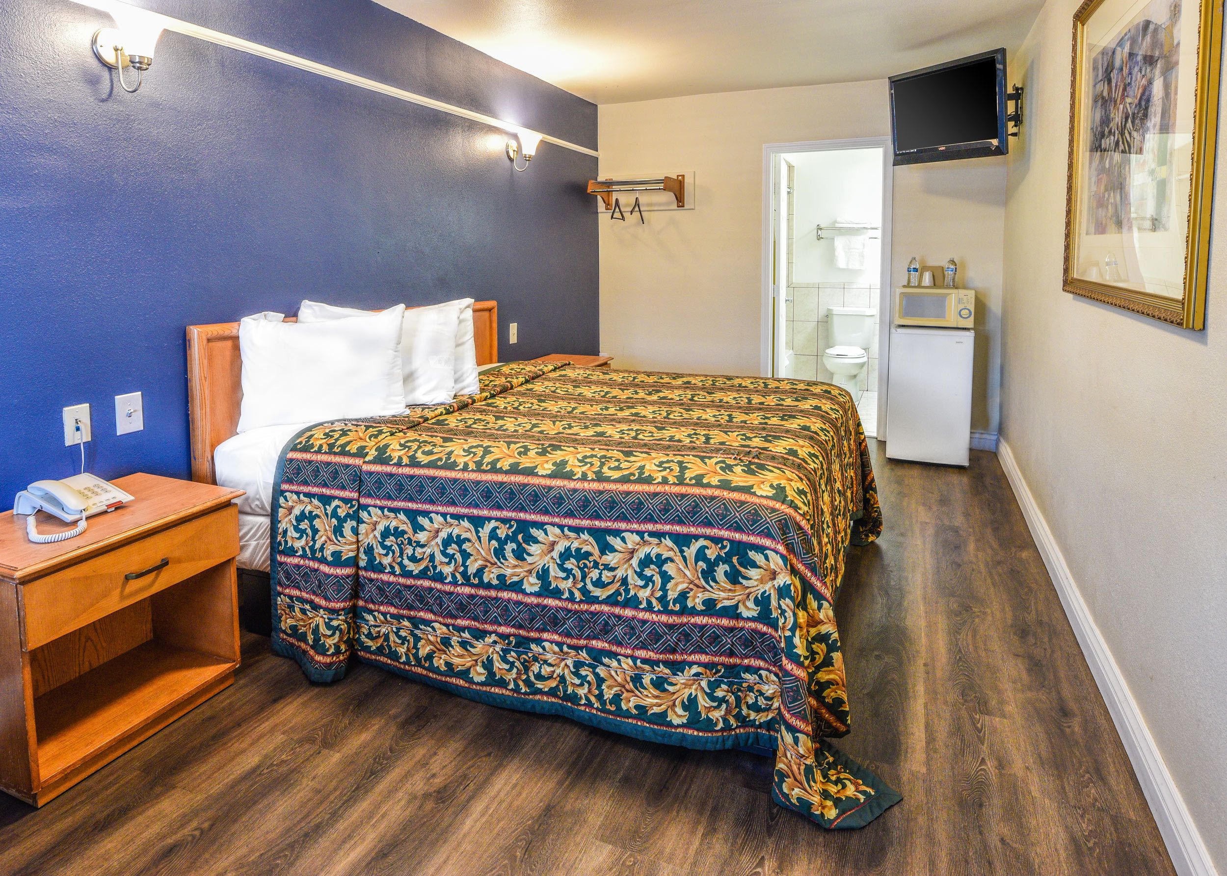 A hotel room with a wooden bed, a bedside table with a phone, a blue wall, a wooden floor, and a door leading to a bathroom.