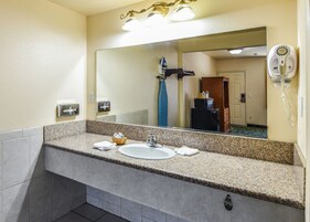 Casa de banho de hotel com bancada de granito, lavatório, espelho, dispensador de toalhas e secador de cabelo.