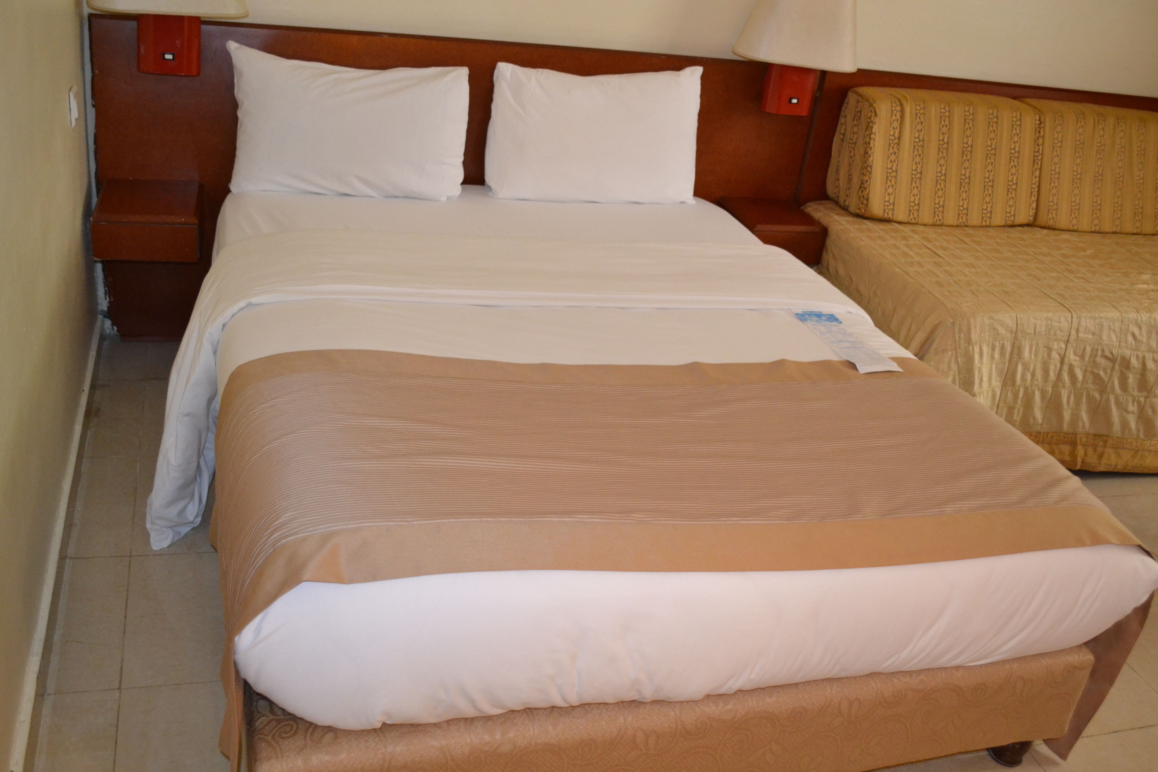 Quarto de hotel com uma cama grande, uma cama menor e uma mesa de cabeceira com abajur.