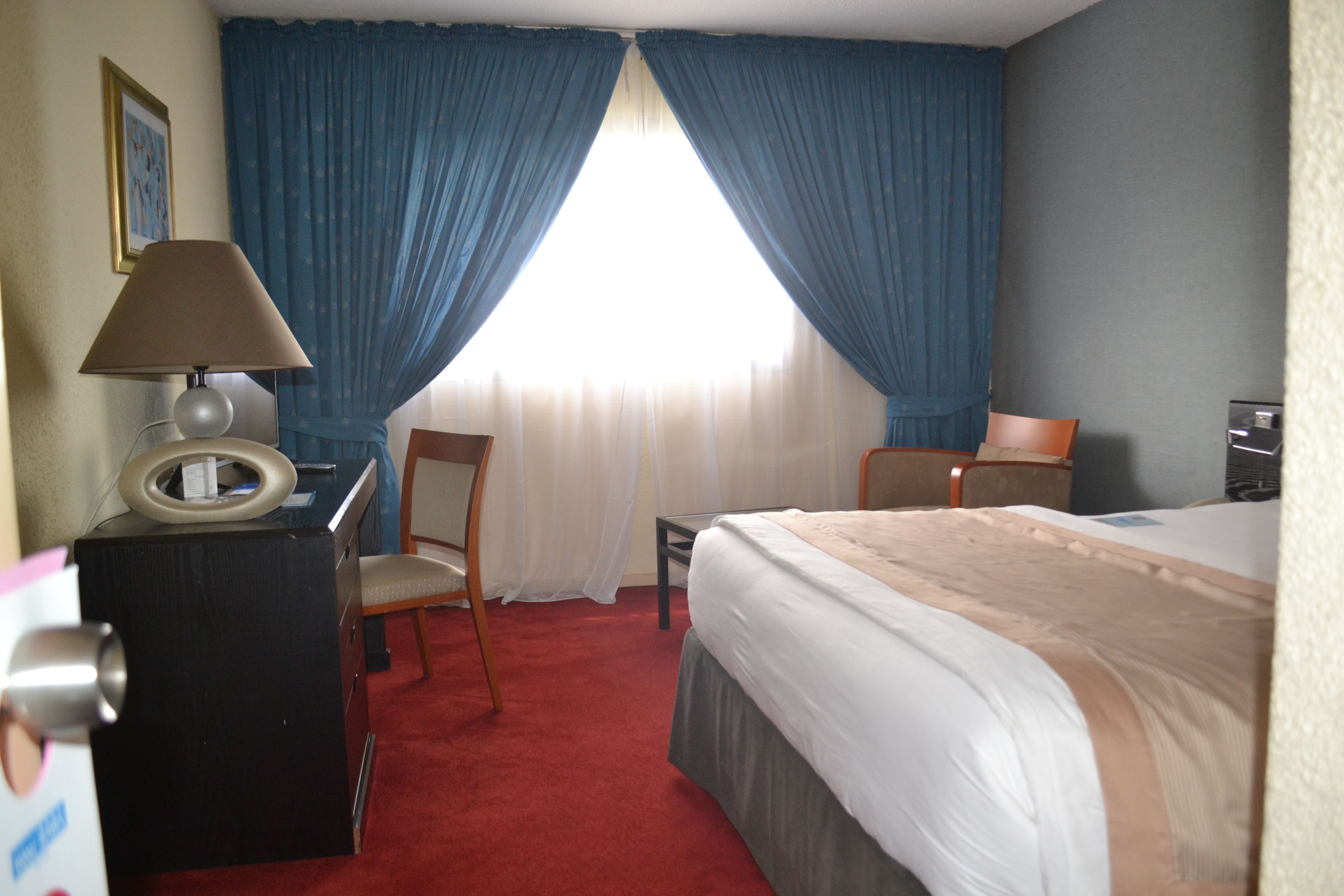 A hotel room with a bed, two chairs, a desk with a lamp, and a window with blue curtains.