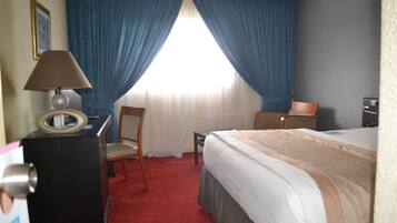 A hotel room with a bed, two chairs, a desk with a lamp, and a window with blue curtains.