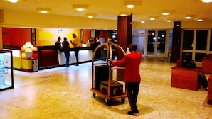 A hotel lobby with a reception desk, a luggage cart, and a television screen displaying a model's image. - Hôtel SAWA (Douala)