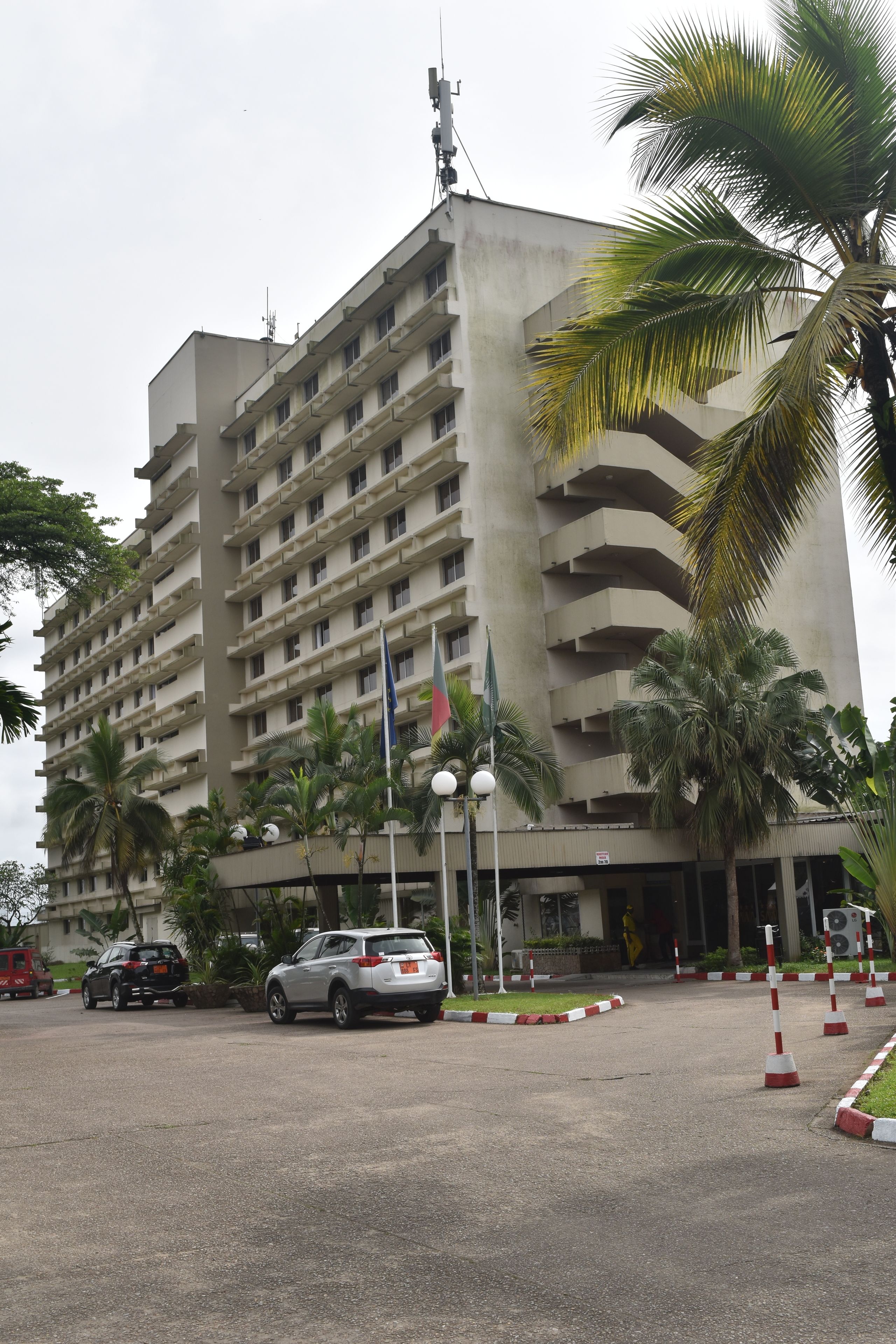 Un hôtel à plusieurs étages, doté d’une entrée principale et entouré de palmiers et de voitures garées.