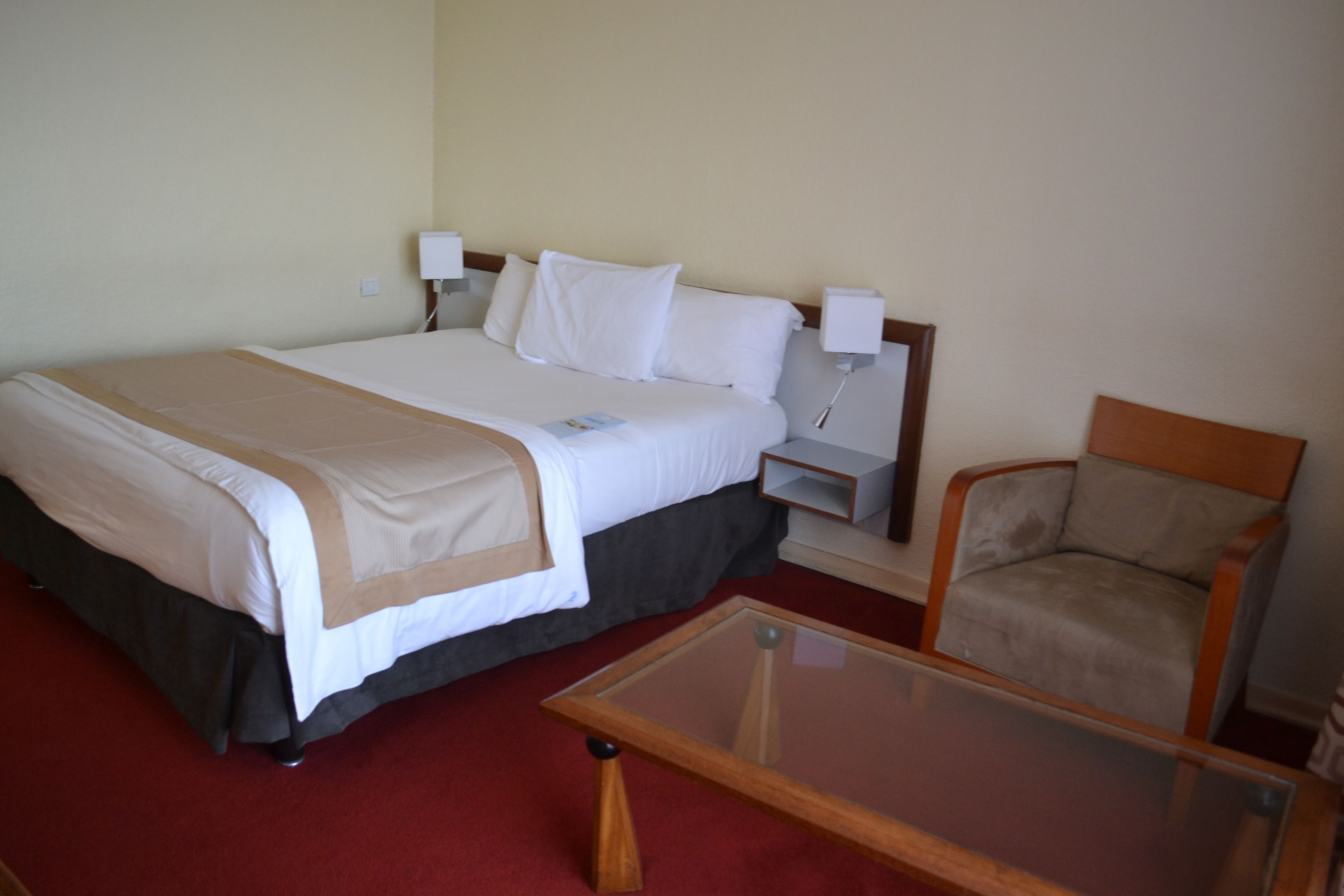 A hotel room with a bed, bedside tables, a chair, and a glass-top coffee table.