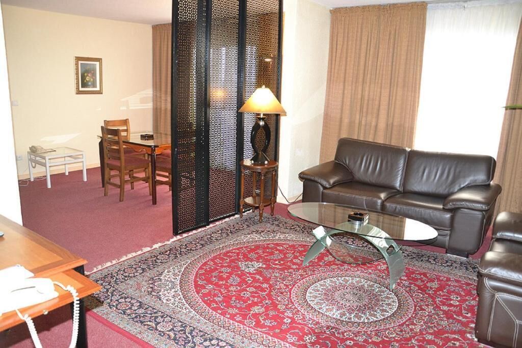 A living room with a leather sofa, a glass coffee table, a patterned rug, a dining table with chairs, and a decorative partition.