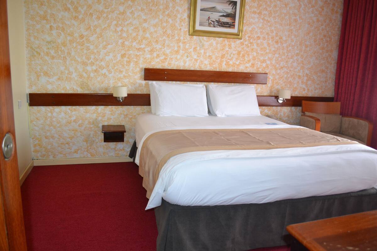 Ein Hotelzimmer mit einem Bett, einem Holzkopfteil, zwei Nachttischen mit Lampen, einem roten Teppich und einer Wand mit Muster.